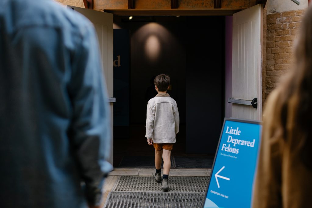 A child walking through an open doorway with a sign outside with the writing, 'Little Depraved Felons'